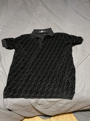 Svart Fendi polotopp med monogram - Säljer en svart polotopp från Fendi med ikoniskt monogrammönster över hela plagget. Klassisk krage och korta ärmar ger en clean look. Perfekt för dig som gillar exklusiva märken och vill sticka ut med stil.