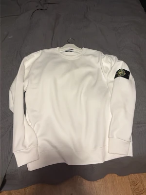 Vit sweatshirt från Stone Island - Säljer en stilren vit sweatshirt från Stone Island med klassisk rund hals och ribbade muddar. Tröjan har den ikoniska Stone Island-loggan på vänster ärm. Perfekt för dig som gillar minimalistisk och trendig streetwear.