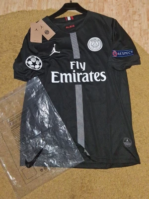 Svart PSG matchtröja från Jordan (ÄKTA) - Snygg svart PSG matchtröja i samarbete med Jordan. Tröjan har vita detaljer, PSG-logga på bröstet, Champions League-märke på ärmen och Fly Emirates-tryck på magen. Tillverkad i lätt och ventilerande material, perfekt för fotboll eller träning. Storleken passar också för M eller även S om du är lång. Säljer pga att den inte passar. Har tags också.