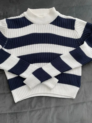 Randig stickad tröja i blått och vitt - Säljer en ribbstickad tröja med breda marinblå och vita ränder. Tröjan har hög hals och långa ärmar, perfekt för dig som gillar en klassisk och stilren look. Materialet är mjukt och stretchigt för extra komfort.