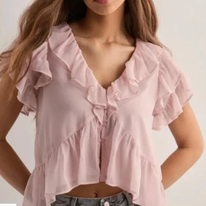 Ljusrosa volangblus från Nelly - Supersöt ljusrosa blus med v-ringning och volangdetaljer längs halsringningen och ärmarna. Croppad modell med korta ärmar och knappar framtill. Perfekt för en trendig och feminin look.