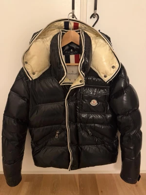 Moncler Dunjacka - Snygg svart dunjacka från Moncler med glansig finish, avtagbar huva och krämvit insida. Jackan har dragkedja, bröstficka med Moncler-logga, två sidofickor med dragkedja och detaljer i vitt och rött på insidan av kragen. Perfekt för kalla vinterdagar. Cartoon sitter löst men finns kvar. Storlek 3 passar s/m