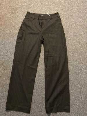 Gråa kostymbyxor med smal rand - Säljer ett par  gråa kostymbyxor med vita nålrandiga ränder. Byxorna har raka ben, hällor för skärp och klassisk knapp- och dragkedjestängning. Perfekta för en clean och stilren look. Byxan är lite liten i storlek, så man tänker på det. 