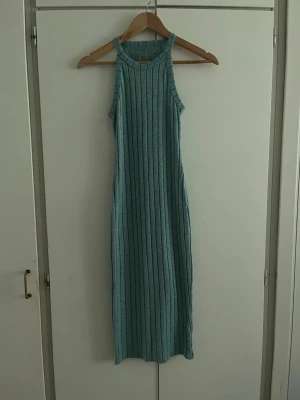 Ljusblå ribbad långklänning - Säljer en ljusblå ribbad långklänning med hög halsringning och ärmlös design. Klänningen har en smickrande rak passform och är tillverkad i ett mjukt stickat material som känns bekvämt mot huden. Perfekt för dig som gillar enkel men snygg stil.