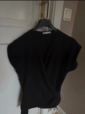 Svart blus - Snygg svart omlottopp från Zara med korta ärmar och v-ringad hals. Toppen har en avslappnad passform och är perfekt för att skapa en stilren look. Tillverkad i ett mjukt material som känns skönt mot huden.
