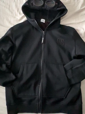 Svart hoodie med glasögon från C.P. Company - Säljer en svart hoodie från C.P. Company med dragkedja och ikoniska glasögon i huvan. Kan gå ner i pris vid snabb affär .