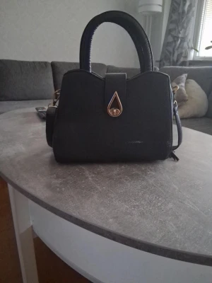 Svart handväska med gulddetalj - Stilren svart handväska med unik form och guldfärgad droppformad låsdetalj framtill. Väskan har två rejäla handtag och avtagbar axelrem. Invändigt finns flera fack och dragkedja för smart organisering. Passar perfekt till dig som gillar klassisk men modern design.