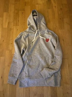 Grå Comme des Garçons Play hoodie - Snygg grå hoodie från Comme des Garçons Play med det ikoniska röda hjärtat med ögon broderat på bröstet. Hoodien har huva med vita snören och en stor magficka. Tillverkad i mjuk bomullsblandning, perfekt för en chill och avslappnad stil. Säljer den pga den är för liten för mig.