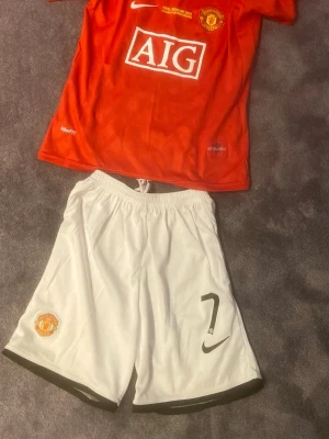 Manchester United Ronaldo matchställ - Säljer ett Manchester United matchställ med röd tröja och vita shorts. Tröjan har AIG-logga, klubbmärke och Ronaldo 7 på ryggen. Shortsen har nummer 7 och klubbmärke. Tillverkat i lätt och ventilerande material, perfekt för fotbollsträning eller match. Storlek 145-155 barn (26 Nike)