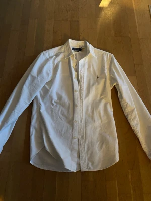 Vit skjorta från Polo Ralph Lauren - Klassisk vit skjorta från Polo Ralph Lauren med broderad logga på bröstet. Slim fit-modell med långa ärmar och knappar framtill. Tillverkad i mjuk bomull som känns skön mot huden. Passar M och S.