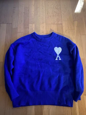 Blå stickad tröja från Ami - Snygg blå stickad tröja från Ami med rund halsringning och ribbade muddar. På bröstet finns ett vitt hjärta med bokstaven A under, vilket är ett signaturmotiv från märket. Passar både M och L