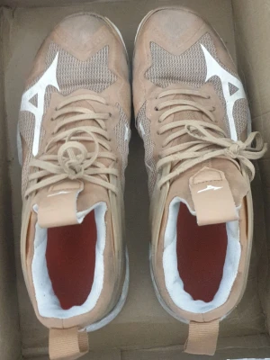 Beige sneakers från Mizuno - Snygga beige sneakers från Mizuno med vita detaljer och meshpaneler för extra ventilation. Skorna har högt skaft, vita sulor och röda innersulor. Perfekta för dig som vill ha en sportig och trendig look.