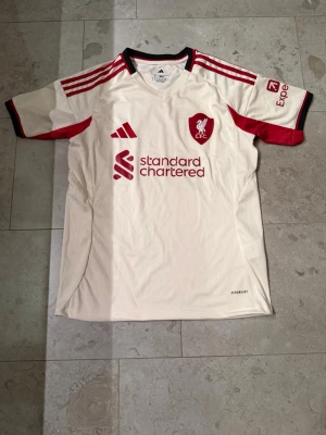 Liverpool bortatröja Adidas vit/röd - Liverpool FC bortatröja från Adidas i vitt med röda detaljer och klubbmärke på bröstet. Tröjan har korta ärmar, svarta och röda ränder på axlarna samt sponsorlogga framtill. Helt ny med lappar