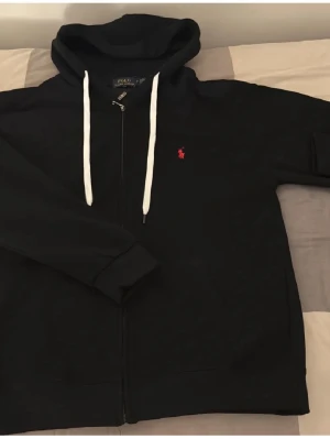 Svart hoodie från Polo Ralph Lauren - Svart hoodie från Polo Ralph Lauren med dragkedja, vit dragsko och röd broderad logga på bröstet. Klassisk design med huva och mjukt material som känns skönt mot huden. Perfekt för en avslappnad och stilren look.