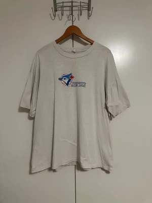 Toronto Blue Jays vit t-shirt - Vit t-shirt med Toronto Blue Jays-logga framtill och stor, färgglad grafik med lagets maskot på ryggen. Klassisk rund hals och korta ärmar. Trycket har blå och röda detaljer som sticker ut mot den ljusa bakgrunden. Perfekt för dig som gillar sportig streetstyle.