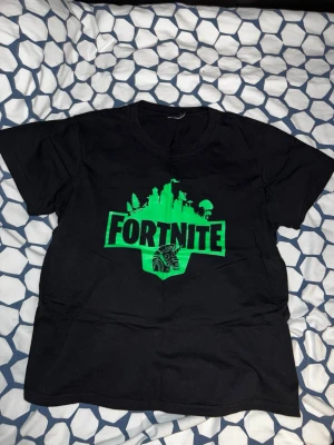 Svart Fortnite t-shirt med tryck - Cool svart t-shirt med stort grönt Fortnite-tryck på bröstet. Klassisk rund hals och korta ärmar. Perfekt för dig som gillar gaming och vill visa din stil. Tillverkad i mjuk bomull för skön känsla hela dagen.