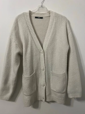 Vit stickad kofta från Bik Bok - Mysig vit stickad kofta från Bik Bok med djup V-ringning och knäppning framtill. Koftan har två stora fickor fram och ribbade muddar vid ärmslut och nederkant. Perfekt för lager på lager och chill stil.