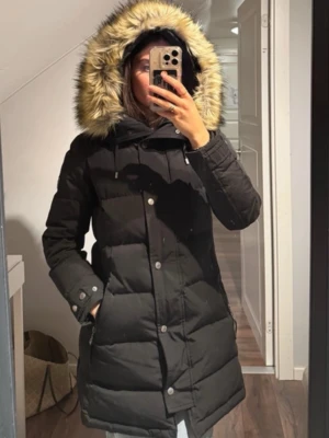 Hollies Dunjacka Subway  - Säljer en svart lång dunjacka med huva och fuskpälsdetalj. Jackan har dragkedja och knappar framtill, fickor med dragkedja och ribbade muddar vid ärmsluten. Puffig modell som håller dig varm under kalla dagar. Märke syns på ärmen.