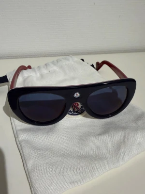 Moncler blå solglasögon - Snygga solglasögon från Moncler med blå bågar och mörka glas. Klassisk oval form med en liten logotyp framtill. Skalmarna är röda och glasögonen kommer med en vit tygpåse med Moncler-logga. Perfekta för dig som vill ha en cool accessoar. Köpta från Vestaire Collective. 