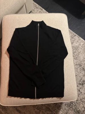 Svart ziptröja med hög krage  - Stilren svart ziptröja med hög krage från Lindbergh och hel dragkedja framtill. Tröjan har långa ärmar och är tillverkad av Australian Merino Wool som passar perfekt till lager-på-lager. Enkel och clean design utan synliga loggor eller detaljer. Org. pris 1699kr 