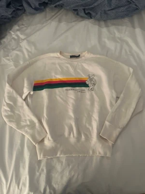 Vit Polo Ralph Lauren sweatshirt med björn - Vit vintage sweatshirt från Polo Ralph Lauren med färgglada ränder i grönt, gult och rött över bröstet samt en ikonisk björn-illustration. Destressed rund halsringning och långa ärmar. 