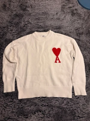 Vit stickad tröja från Ami Paris - Vit stickad tröja från Ami Paris med röd hjärtlogga och bokstaven A på bröstet. Tröjan har rund halsringning, ribbade muddar och nederkant. Perfekt för dig som gillar stilrena plagg med en twist.