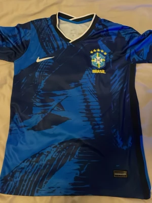 Blå Brasilien Nike fotbollströja - Snygg blå fotbollströja från Nike med Brasilien-emblem och fem stjärnor på bröstet. Tröjan har ett unikt abstrakt mönster i olika blå nyanser, vit Nike-logga och gul text. Tillverkad i lätt och ventilerande material, perfekt för träning eller match.