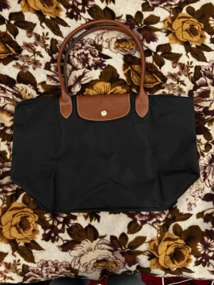 Svart Longchamp Le Pliage handväska - Klassisk svart Le Pliage handväska från Longchamp med bruna läderhandtag och lock. Väskan är tillverkad i slitstark nylon med detaljer i brunt läder och har en guldknapp framtill. Perfekt storlek för att få plats med allt du behöver och enkel att bära över axeln.