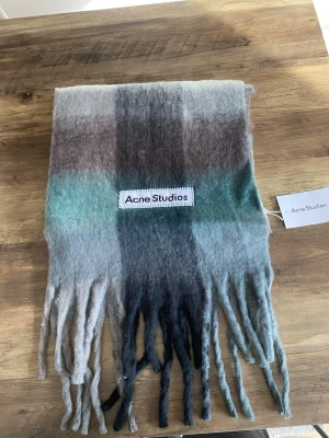 Rutig halsduk från Acne Studios - Mjuk och fluffig halsduk från Acne Studios i rutigt mönster med nyanser av grått, brunt, blått och grönt. Stor logotyp framtill och långa fransar i ändarna. Perfekt accessoar för att lyfta din outfit med en trendig touch.