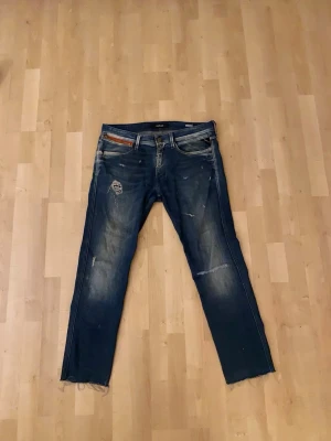 Blåa jeans från Replay - Säljer ett par blå replay anbass jeans med slitningar och avklippta benslut pga av för långa. Jeansen har klassisk femficksdesign och kontrastsömmar. Perfekta för en avslappnad och trendig look. Säljer då det är för små