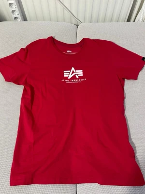 Röd Alpha Industries t-shirt - Snygg röd t-shirt från Alpha Industries med vit logga och text på bröstet. Använd fåtal gånger samt i fint skick. 