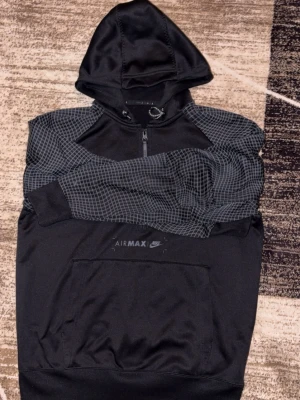  Nike Air Max hoodie  - Snygg svart hoodie från Nike Air Max med halv dragkedja och huva. Ärmar och axlar har ett rutigt mönster i grått som ger en sportig vibe. Stor magficka framtill och diskret Air Max-logga på bröstet. Perfekt för dig som gillar streetwear och sportig stil.