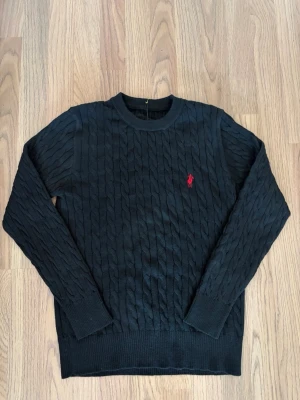 Svart tröja - Kabelstickad svart tröja från med häst på med rund halsringning och den klassiska röda logotypen broderad på bröstet. Tröjan har långa ärmar och ribbade muddar vid ärmslut och nederkant. Perfekt för en stilren och tidlös look.