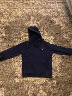 Mörkblå hoodie från Lyle & Scott - Mörkblå hoodie från Lyle & Scott med klassisk gul logga på bröstet. Hoodien har en stor känguruficka framtill och justerbar huva med dragsko. Tillverkad i mjukt bomullsmaterial, perfekt för en avslappnad och stilren look.