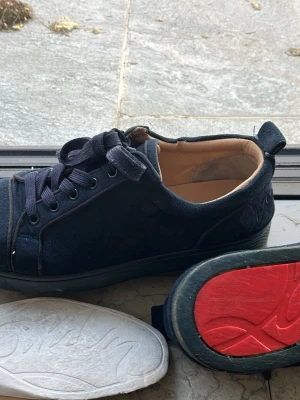 Blåa sneakers från Christian Louboutin - Snygga blåa sneakers från Christian Louboutin i mocka med klassiska röda sulor. Skorna har blå skosnören, metallöljetter och låg profil. Perfekta för dig som vill ha en exklusiv och stilren look med en ikonisk touch.
