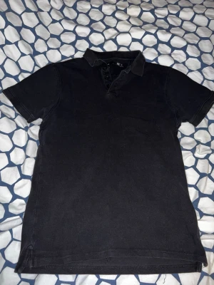 Svart pikétröja med krage - Klassisk svart pikétröja med krage och knappar framtill. Kortärmad modell i mjukt bomullsmaterial som passar perfekt till jeans eller shorts. Enkel och stilren design utan tryck eller mönster.