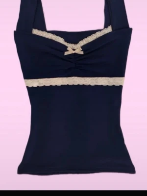 Mörkblå linnetopp med spets och rosett - Supersöt mörkblå linnetopp med breda axelband, dekorerad med vit spetsdetaljer och en liten rosett framtill. Toppen har en figurnära passform och är perfekt för att skapa en söt och trendig look. Är inte riktigt min stil🤍