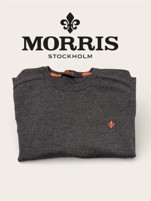 Mörkgrå stickad tröja Morris - Mörkgrå stickad tröja från Morris med rund halsringning och diskret orange logga broderad på bröstet. Tröjan har långa ärmar och ribbade muddar vid ärmslut och nederkant. Perfekt för lager på lager och stilren look.