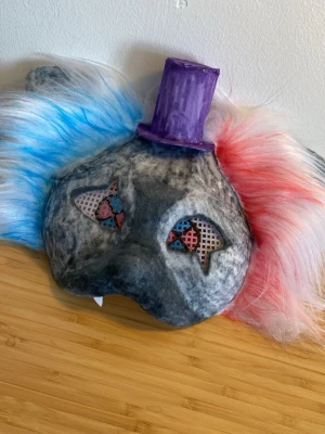 Clownmask med hatt och färgat hår - Unik clownmask med fluffigt syntethår i blått och rött, grå bas och fastsydd lila hatt. Masken har rutiga detaljer vid ögonen och elastiskt band för enkel användning. Perfekt till maskerad eller cosplay för dig som vill sticka ut.