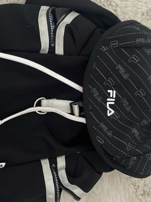 Svart ryggsäck från FILA med silverdetaljer - Snygg svart ryggsäck från FILA med vita och silverfärgade detaljer. Ryggsäcken har ett stort huvudfack, dragkedjor med silverband och ett runt ytterfack med FILA-logga i vitt. Köpt för 500 säljer för 200