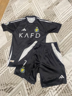 Al Nassr svart fotbollsset Adidas - Snyggt svart fotbollsset från Al Nassr med matchtröja och shorts. Tröjan har vita detaljer, klubbmärke och KAFD-tryck på bröstet. Shortsen har nummer 7 och klubbmärke. Tillverkat i lätt och ventilerande material, perfekt för träning eller match. Jätte fin Ronaldo tröja, titta på bilden shortsen e lite förstörd men absolut inga problem. Skulle säga passar 14 åringar