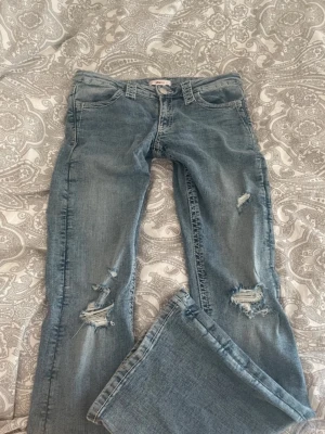 Ljusblå slitna bootcut jeans - Säljer ett par ljusblå bootcut jeans med slitningar på knä och lår. Jeansen har låg midja och klassisk femficksmodell. Perfekta för en avslappnad och trendig look. Materialet är denim i bomull. Passar dig som gillar 00-talsstil.