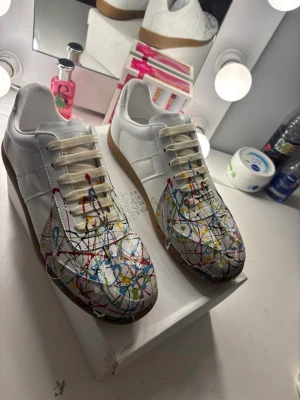 Maison Margiela sneakers med färgstänk - Unika vita sneakers från Maison Margiela med färgstänk i rött, blått, gult och svart över tå och sidor. Skorna har klassisk låg siluett, beige snören och gummisula. Tillverkade i läder och textil för en clean men kreativ vibe.