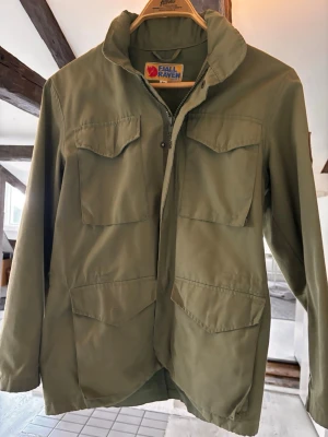 Olivgrön vindjacka från Fjällräven - Snygg olivgrön vindjacka från Fjällräven med fyra stora fickor framtill och hög krage. Jackan har dragkedja och tryckknappar, samt klassisk outdoor-look. Tillverkad i slitstarkt material som passar perfekt för blåsiga dagar.