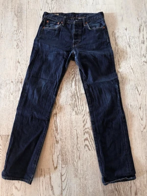 Mörkblå Levi's 501 - Säljer ett par klassiska mörkblå jeans från Levi's med rak passform. Jeansen har fem fickor, orange sömmar och en brun läderpatch bak i midjan. Tillverkade i slitstark denim, perfekta för en avslappnad och tidlös stil.