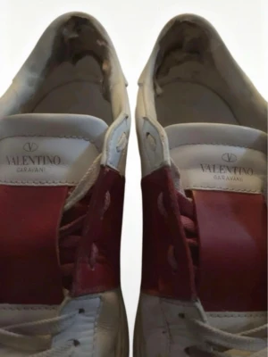 Valentino Garavani vita sneakers - Snygga vita sneakers från Valentino Garavani med bred röd remsa över sidan. Skorna har klassisk låg profil, vita snören och detaljerade nitar på sulan. Tillverkade i läder för en exklusiv känsla och stilren look.