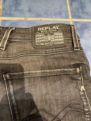 Svarta Replay jeans med slitning - Säljer ett par svarta jeans från Replay med klassisk femficksmodell och tydliga kontrastsömmar. Jeansen har en snygg tvättad look och Replay-logga på bakfickan. Perfekta för dig som gillar streetwear och vill ha ett par jeans med karaktär.