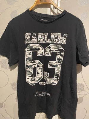 Svart Harlem 63 t-shirt från Primark - Cool svart t-shirt från Primark med stort tryck på bröstet: 'HARLEM 63' där siffrorna har ett grått camouflage-mönster. Klassisk rund hals och korta ärmar. Perfekt för en streetwear-inspirerad look.