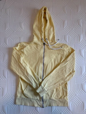Ljusgul hoodie Lager157 - Ljusgul hoodie med dragkedja framtill och justerbar huva med vita snören. Hoodien har långa ärmar, ribbade muddar och två fickor framtill. 