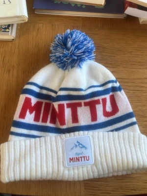 Vit och blå Minttu mössa med tofs - Säljer en vit stickad mössa från Minttu med blåa ränder och röd text framtill. Mössan har en stor blåvit tofs på toppen och vikt kant med Minttu-märke. Perfekt för kalla dagar och snygg till vinterlooken.